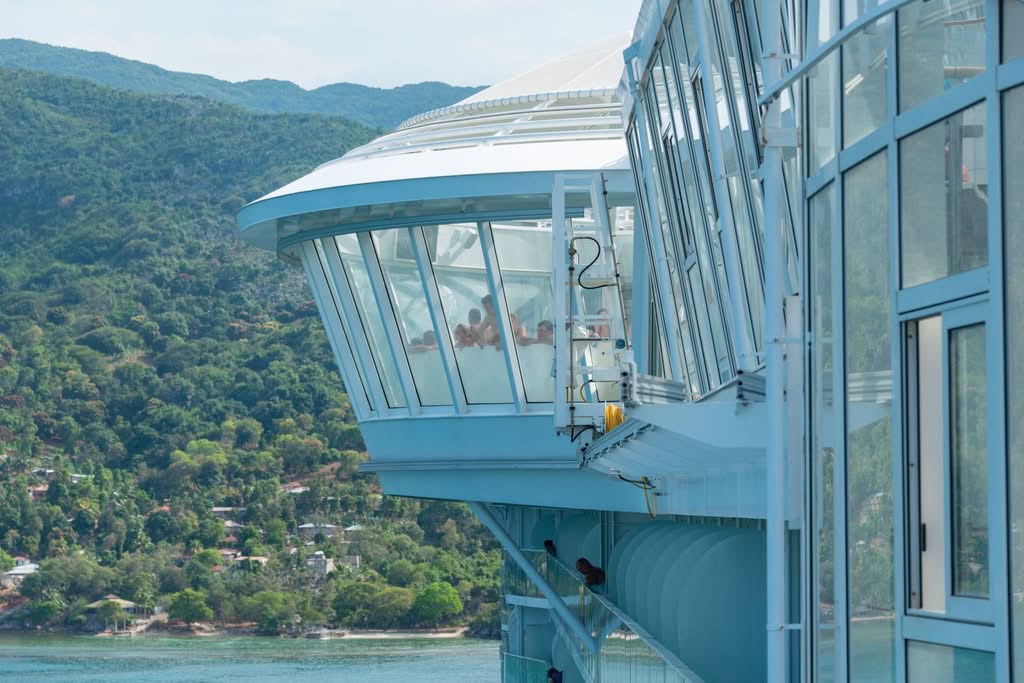 A modern cruise ship deck with glass enclosure overlooking a lush coastal landscape