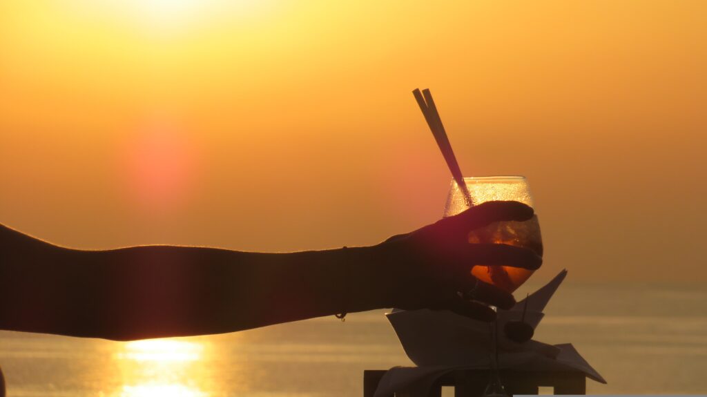 A hand raising a cocktail glass silhouetted against a golden sunset over the sea